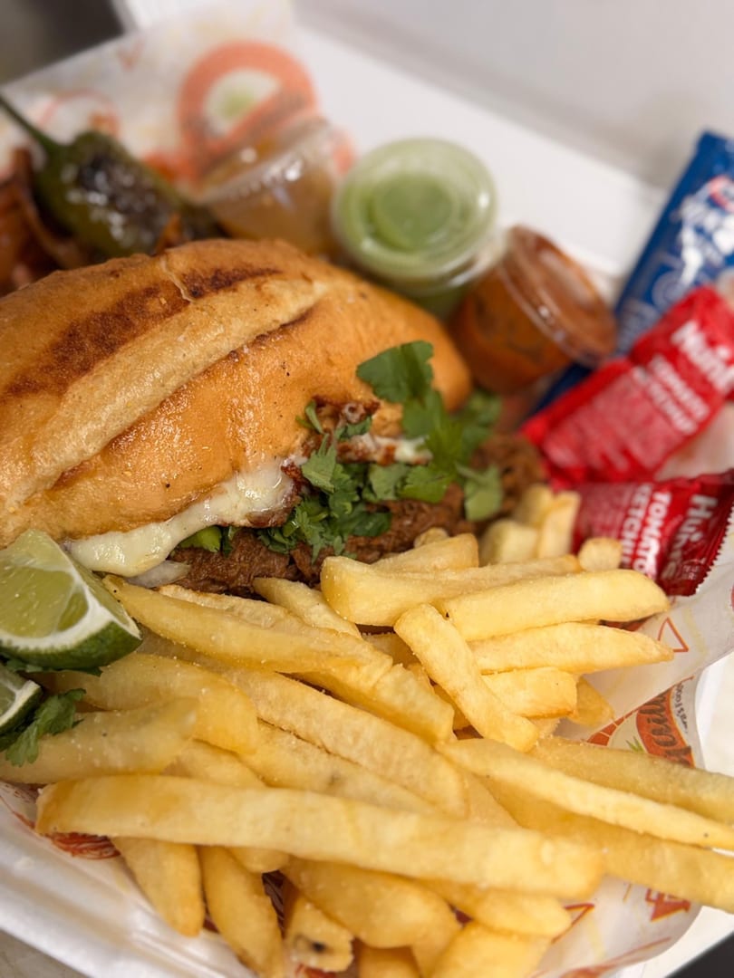 A close-up of a torta sandwich with melted cheese, cilantro, lime, fries, and sauces.